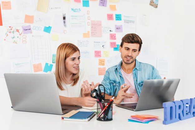 Two colleagues discussing work with a laptop and sticky notes on the wall.