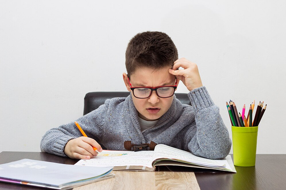 A boy with glasses studies intensely at a desk filled with books and stationery.