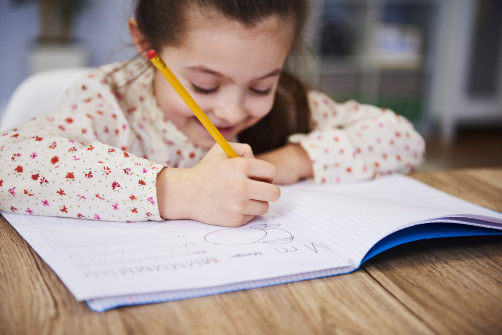 A young girl focused on writing in a notebook with a pencil.