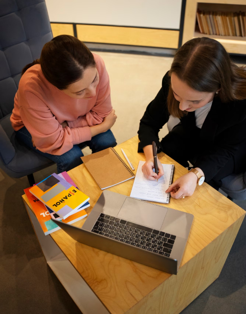 Two women working together at a table with notebooks and a laptop.