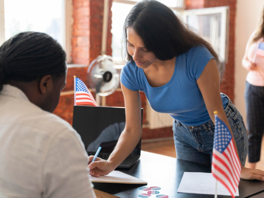 A woman assisting a man with paperwork at a desk adorned with small American flags.